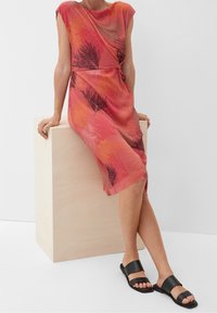 Vestido con estampado rojo y naranja, de mangas cortas, con detalles drapeados y una abertura lateral, combinado con sandalias planas negras sobre un fondo neutro.