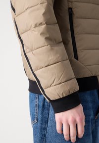 Casaco puffer bege com textura acolchoada, acabamento preto nas mangas e bolso com fecho, combinado com jeans de ganga azuis.