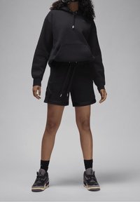 Felpa nera con tasca frontale e coulisse, abbinata a pantaloni corti neri con tasche laterali. Sneakers nere completano l'outfit.