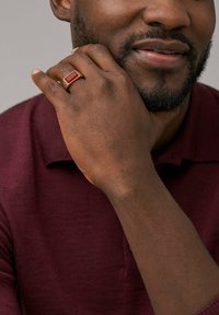 Guldring med en rektangulær rød sten, holdt op mod en maroon polo t-shirt. Ringen har en glat overflade og rene linjer.