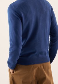 Man som bär en blå långärmad tröja och bruna byxor med händerna i fickorna, sett bakifrån mot en neutral bakgrund.