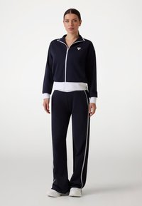 Marineblaue Trainingsanzug mit weißen Akzenten, bestehend aus einer Reißverschlussjacke und passenden Hosen, beide aus weichem Material. Die Jacke hat ein dreieckiges Logo.