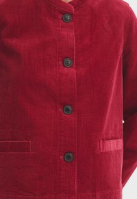 Rode corduroy jas met een V-hals, vijf zwarte knopen en een voorzak met verticale ribbelstructuur. Zachte textuur en gestructureerd ontwerp.