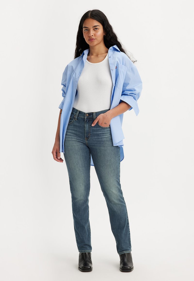 Helles Blaues Hemd über einem weißen Tanktop, kombiniert mit dunkelblauen Jeans und schwarzen Stiefeletten, die einen lockeren Schnitt und einen lässigen Stil zeigen.