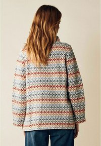 Kvinna med långt vågigt hår som bär en flerfärgad stickad tröja och blå jeans, vänd bort från kameran mot en neutral bakgrund.