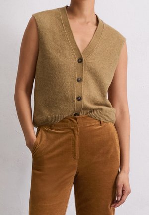 Henkilö, jolla on yllään hihaton, napitettava ruskea neuleliivi sekä korkea vyötäröiset, ruosteenväriset puuvillahousut, kädet rentoina sivuilla.