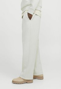 Pantaloni de trening gri deschis, cu o croială relaxată, prevăzuți cu buzunare laterale și manșete cu nervuri, asortați cu încălțăminte bej. Textură netedă și design minimalist.