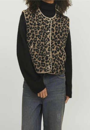 Person iført leopardmønstret knappet vest over sort rullekrave og blå jeans, stående med den ene hånd ved siden.