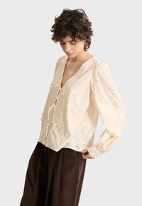 Cremfarbene, bestickte Bluse mit Bindeschluss, V-Ausschnitt, langen Puffärmel und geometrischen Cut-out-Mustern. Getragen mit braunen Cordhosen.