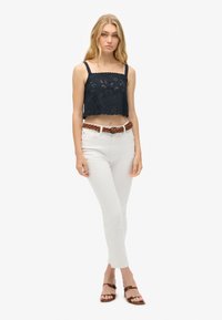 Marineblauwe kanten crop top met een gegolfde rand, gecombineerd met witte skinny jeans en een gevlochten bruine riem. Bruine sandalen maken de outfit compleet.