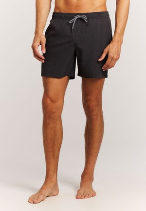 Homme portant un short de bain noir avec une taille élastique et un cordon noir et blanc, debout pieds nus sur un fond uni.