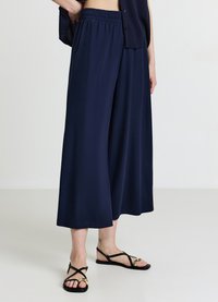 Pantalon large bleu marine en tissu doux avec une taille élastique, associé à des sandales plates noires avec des accents dorés.