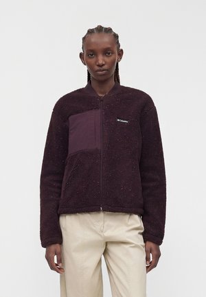 Jeune femme aux cheveux tressés portant une veste polaire violet foncé avec une poche avant et un pantalon beige, debout devant un fond uni.