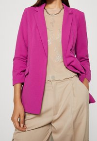 Fuchsiablazer med skräddarsydd design, strukturerade axlar och långa ärmar, bärs över en beige topp med silverdetaljer.
