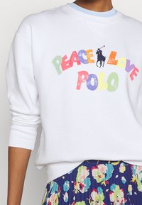 Bílá mikina se grafickým nápisem "Peace Love Polo" v multibarevných písmenech. Má žebrované manžety a kulatý výstřih. Doplňuje ji květinová sukně.