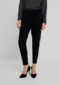 Pantalones de chándal negros hechos de material suave, con cintura ajustable mediante cordones, bolsillos laterales con cremallera y tobillos ajustados. Combinados con tacones negros.