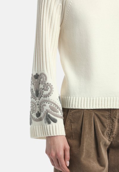 Maglione di colore crema a maglia con texture a coste, caratterizzato da ricami grigi decorativi sulle maniche e un orlo a coste.