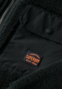 Czarna kurtka z wyściółką z polaru, z czarną panelem z materiału. Posiada pomarańczową i niebieską haftowaną łatę z logo oznaczoną "Superdry Work-Wear".