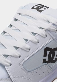 Λευκό αθλητικό παπούτσι με λευκά κορδόνια, μαύρο λογότυπο DC στη πλευρά και τη γλώσσα, δερμάτινα και πλεκτά πάνελ με υφή και ραφές.