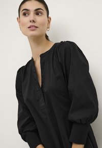 Črna tunika z v-izrezom, napihnjenimi rokavi in rebrastimi manšetami. Tkanina ima gladko, mat teksturo brez vzorcev.