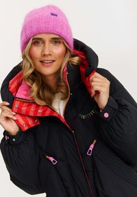 Čierna nadýchaná bunda s ružovým pruhovaným podšívkou, farebným prešívaním a ružovými zipsami; doplnená mäkkou svetloružovou pletenou čiapkou.