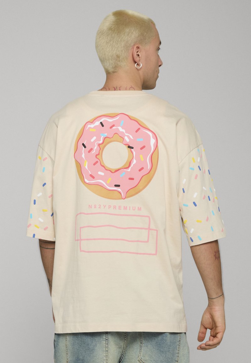 Béžové oversized tričko s velkým růžovým obrázkem donutu na zádech, zdobené barevnými posypy na rukávech a štítkem pod ním.