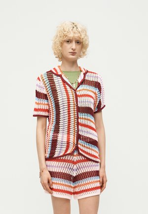 Osoba s kučeravými blond vlasmi oblečená v viacfarebnej otvorene pletenej krátkorukávovej košeli a zodpovedajúcich šortkách, stojaca pred jednoliatym svetlým pozadím.