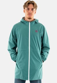 Veste en teal résistante à l'eau avec une fermeture éclair à l'avant, capuche avec cordon de serrage, deux poches latérales et un petit logo circulaire sur la poitrine.