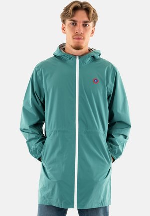 Veste en teal résistante à l'eau avec une fermeture éclair à l'avant, capuche avec cordon de serrage, deux poches latérales et un petit logo circulaire sur la poitrine.