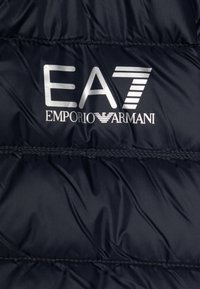 Navy puffer jakna od tkanine s kvadratnom teksturom; sadrži srebrni EA7 logo i tekst "Emporio Armani" u kontrastnim bojama.