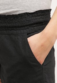 Schwarze Leinenhose mit elastischem Bund, seitlichen Taschen und glatter Textur. Das Design bietet einen lockeren Schnitt und eine schlichte Verarbeitung.