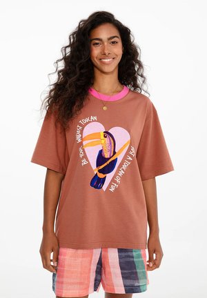Uśmiechnięta kobieta z długimi kręconymi włosami nosi brązowy T-shirt z ilustracją tukana i wielokolorowe, paskowane szorty na białym tle.