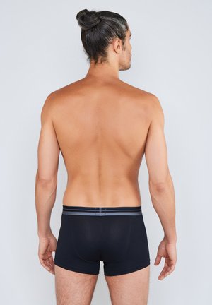 Uomo adulto con capelli scuri raccolti in un ciuffo, visto da dietro, che indossa boxer neri con una fascia in vita grigia su uno sfondo chiaro e uniforme.