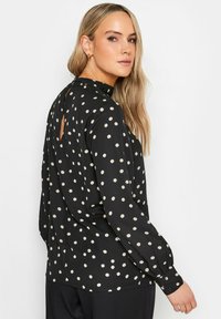 Svart blus med krämfärgade prickar, långa ärmar, hög halsringning och en nyckelhålsdetalj bak. Mjuk tyg med en slät yta.