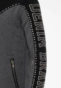 Grauer und schwarzer Sweatshirt mit texturiertem Stoff, Reißverschlusstasche und metallischen Nieten entlang des Ärmels, mit dem auffälligen Text "DENIM BRAND."