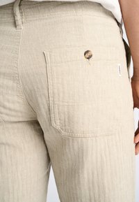 Lichtbeige linnenbroeken met een visgraatpatroon, een achterzak met een knoop en een zijkleurlabel. Gladde textuur en een getailleerde pasvorm.