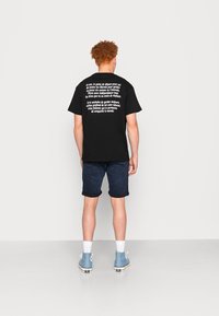 Musta t-paita, jossa on valkoista tekstiä selässä, yhdistetty tummiin farkkushortsisiin. Paidassa on rento malli ja lyhyet hihat. Jalassa on siniset tenniskenkiä.