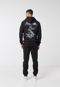 Muž stojí zády, oblečený v černé mikině s kapucí s motivem sovy a bílým textem, černých sportovních kalhotách a bílých teniskách, na jednobarevném pozadí.