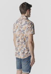 Hombre visto de espaldas que lleva una camisa de manga corta con estampado floral en beige, naranja y azul, combinada con pantalones cortos de mezclilla remangados.