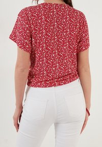 Blouse rouge à motif floral blanc, manches courtes et ourlet arrondi ; associée à un jean blanc avec poches arrière et coupe slim.