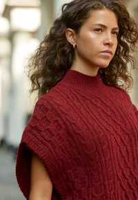 Poncho en maille torsadée de couleur bordeaux profond, avec un col montant et des côtés ouverts. Motif texturé avec une apparence douce, semblable à de la laine.