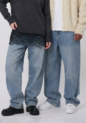 Két ember laza, világoskék farmert visel fekete platformcsizmával és fehér tornacipővel, réteges kötött pulóverekkel párosítva.