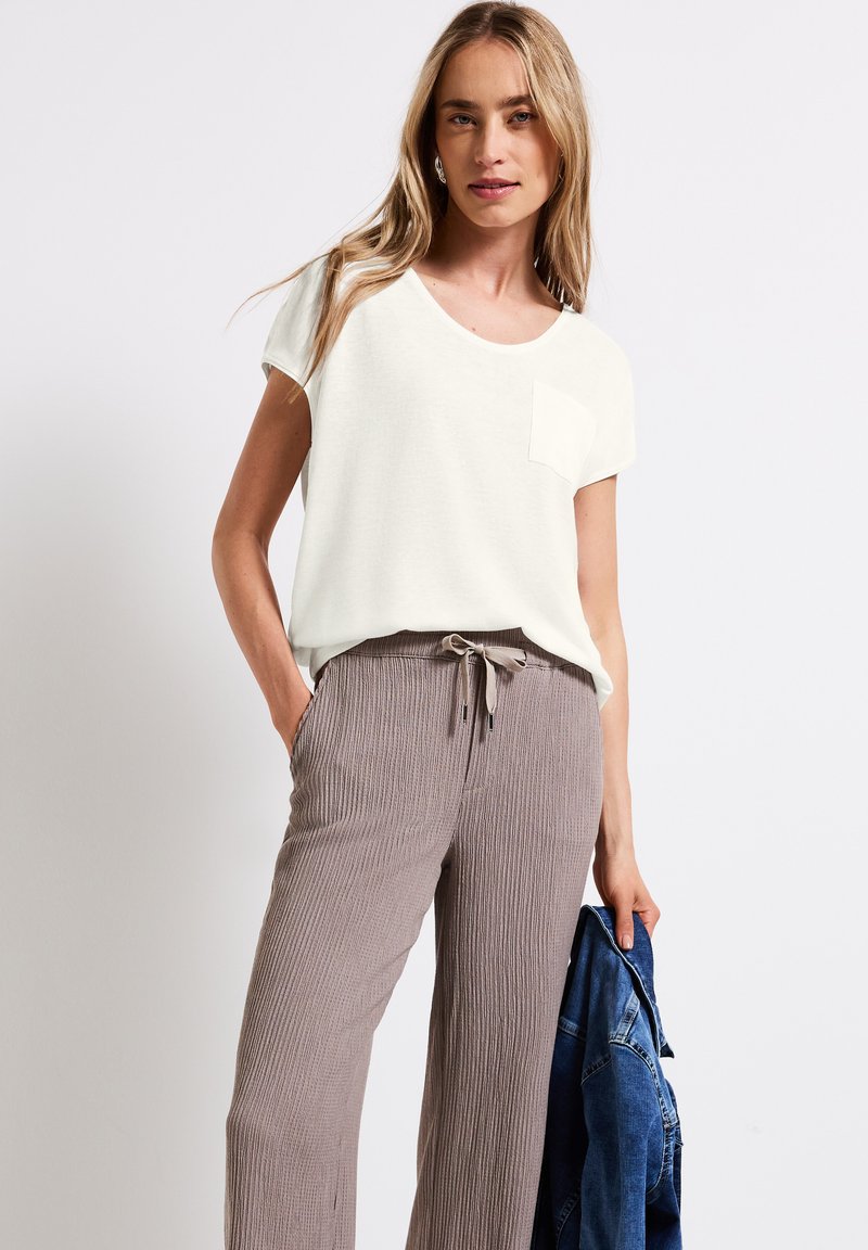 T-shirt crema a maniche corte con taschino, abbinato a pantaloni a coste marrone chiaro con cintura elastica e cordoncino. Giacca di jeans in mano.