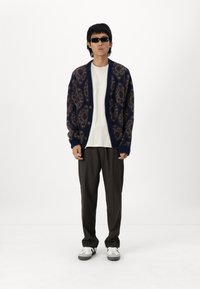 Navy patterned cardigan with brown accents, white T-shirt, dark trousers, and gray shoes with black stripes, worn by a person.