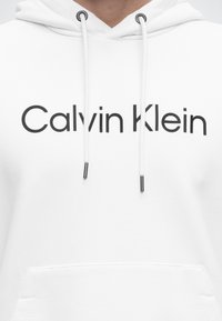 Fehér kapucnis pulóver fekete "Calvin Klein" felirattal a mellrészen, fehér húzózsinórokkal, amelyeken fekete fémvégek találhatók, valamint elöl egy erszénytáska-zsebbel.
