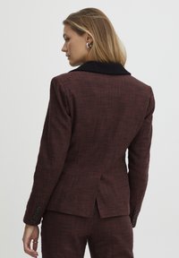Blazer à carreaux marron avec col noir. Tissu texturé, poignet à un bouton, et design avec fente au dos. Coupe ajustée pour un look structuré.