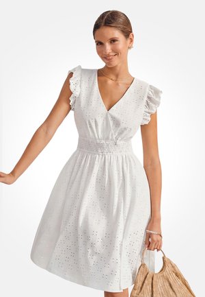 Frau trägt weißes Kleid mit Lochstickerei, Rüschenärmeln und geraffter Taille, hält gewebte Handtasche, lächelt vor weißem Hintergrund.