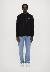 Zwarte zip-up hoodie met koordjes, voorzien van een gele logo op de borst en de mouw. Gepaarde met lichtblauwe jeans en witte sneakers.