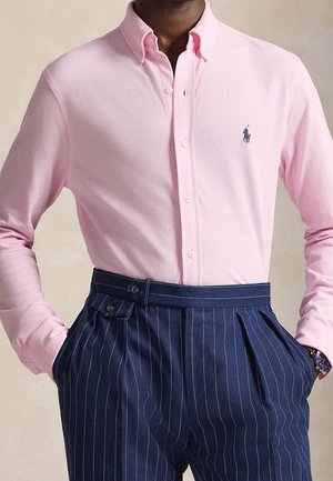 Homme portant une chemise rose clair boutonnée avec un petit logo et un pantalon à pinces bleu marine taille haute, les mains dans les poches.