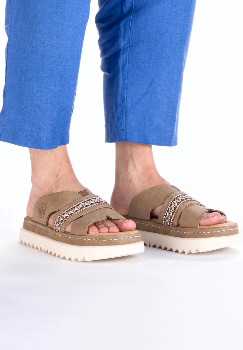 Brown suede slip-on sandals with white accents and textured rubber sole, featuring cross straps and woven detail across the top.